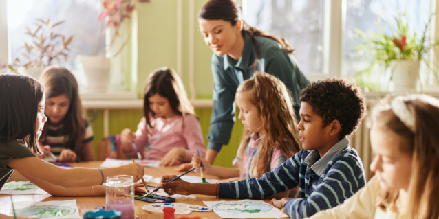 Eine Gruppe Grundschüler sitzt an einem Tisch und malt mit Wasserfarbe und die Lehrerin schaut zu