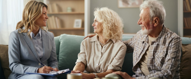 Junge Frau in Anzug sitzt mit altem Paar auf dem Sofa und schreibt auf einem Dokument