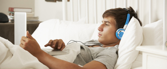 Junge mit Kopfhörern liegt im Bett, mit dem Laptop auf seinem Bauch