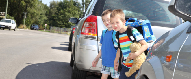 Einschulung: Schulkinder mit Ranzen und Teddbär im Arm zwischen parkenden Autos am Straßenrand