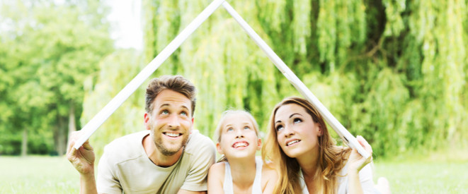 junge Familie liegt lächelnd auf dem Bauch auf dem Rasen und hält ein Dach über sich