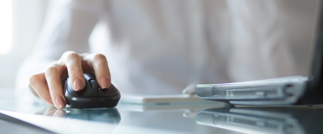 Nahaufnahme: Frauenhand führt Computermaus auf einer Schreibtischoberfläche, daneben ein aufgeklappter Laptop