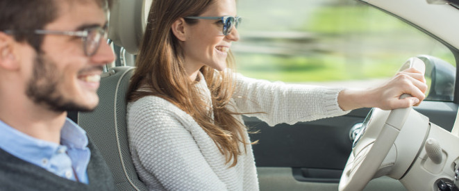 Probefahrt mit Gebrauchtwagen: Junge Frau mit Brille fährt Auto, auf dem Beifahrersitz ein junger Mann mit Brille