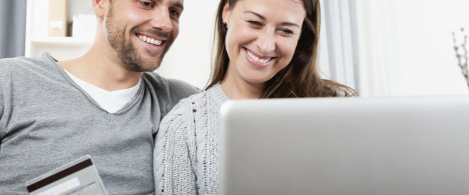junges Paar sitzt lächelnd vor einem Computer, Mann hält eine Kreditkarte in der Hand