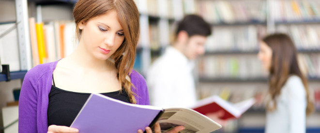 Studentin liest in der Uni-Bibliothek ein Buch, im Hintergrund zwei Studenten