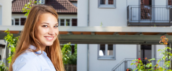 Lächelnde, junge Mietern steht vor einem Mehrfamilienhaus