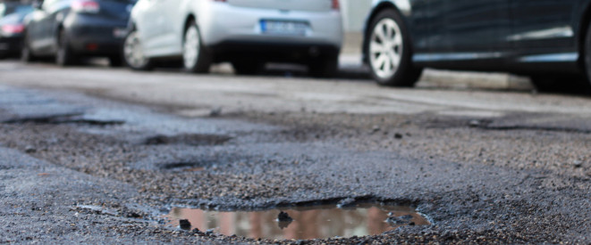 Eine unebene Straße mit Schlaglöchern, an desen Rand parkende Autos stehen.