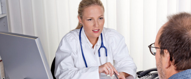 Berufskrankheit: Diese Voraussetzungen gelten. Eine blonde Ärztin sitzt einem Patienten gegenüber.