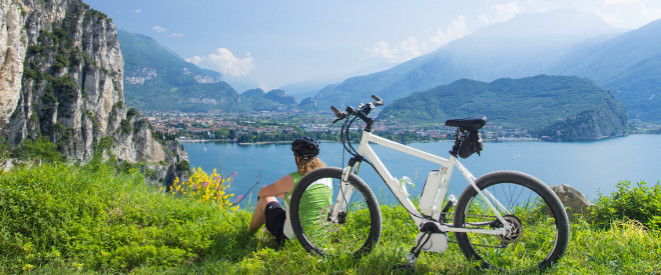 E-Bike nachrüsten: Diese Regelungen sind zu beachten. Eine Frau sitzt in einer Berglandschaft mit Ihrem E-Bike auf einer mit Gras bewachsenen Klippe und blickt einen türkisen See herunter.