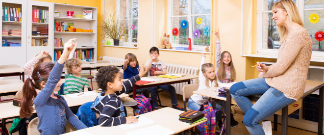 Freistellung vom Unterricht: In diesen Fällen ist sie erlaubt. Eine blonde Lehrerin sitzt auf einem Tisch vor einer kleinen Grundschulklasse.