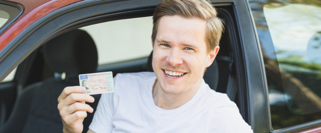 Führerschein auf Probe: Besondere Regeln für Fahranfänger. EIn junger Mann guckt lächelnd aus einem Auto und hält einen Führerschein in seiner Hand.