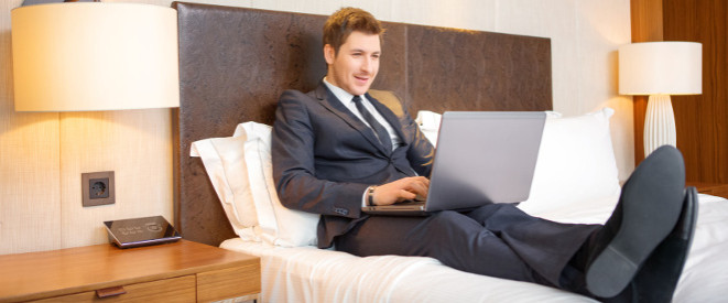 Reisekostenerstattung: Diese Rechte haben Arbeitnehmer. Ein Mann in schwarzem Anzug sitz auf einem Hotelzimmer-Bett und hat einen Laptop auf dem Schoß. 