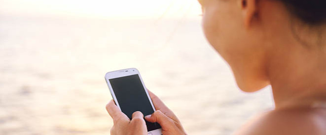 Roaming-Gebühren in der EU: Tipps für den Urlaub 2016. Eine Frau mit Strohhut guckt am Strand auf ihr Smartphone.
