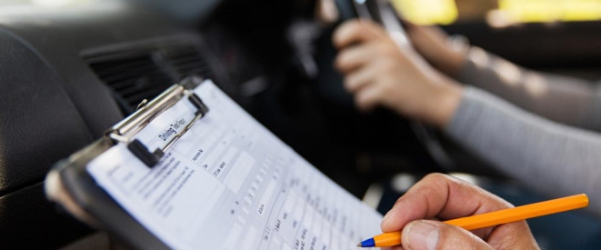 Bei der Fahrprüfung durchgefallen: So geht es weiter. Ein Mann macht Notizen auf einem Klemmbrett, dahinter fässt eine andere Person ans Steuer eines Autos.
