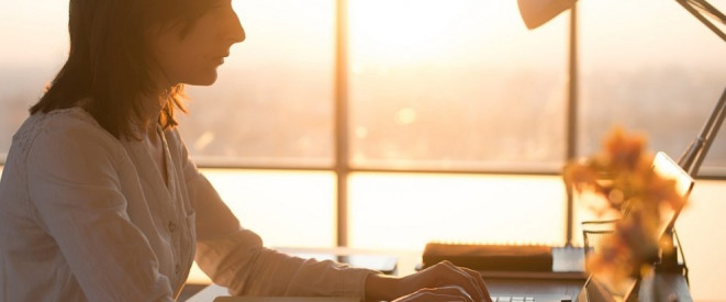 Doppelte Haushaltsführung: Zweitwohnung absetzen – die Regeln. Ein Frau sitzt im Licht des Sonnenuntergangs am Schreibtisch und tippt auf einem Notebook.