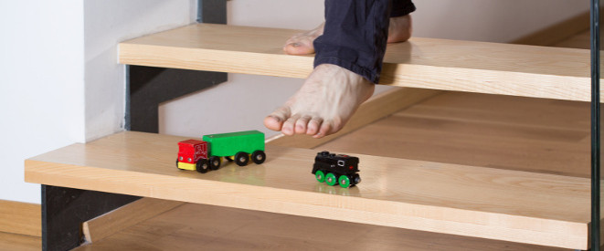 Homeoffice: Unfallversicherung greift nicht immer. Ein nackter Fuß steigt auf eine Holzstufe zwischen zwei Spielzeugautos hinab.