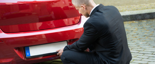 Kennzeichen verloren: Tipps für das Behelfskennzeichen. Ein Mann in scharzem Anzug montiert das hintere Kennzeichen eines roten Autos.