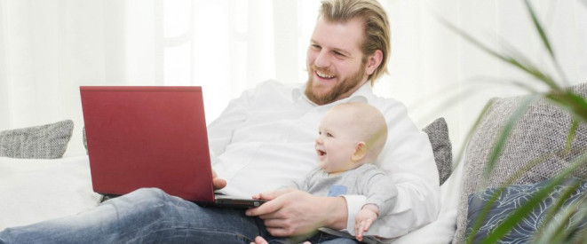 Urlaubsanspruch bei Elternzeit: Die Rechtslage. Ein blonder Mann mit Bart sitzt lachend auf einem Sofa, hält ein Baby im Arm und hat ein rotes Notebook auf dem Schoß.