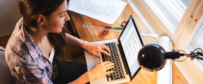 Gehalt und Einkommensgrenze bei Werkstudenten. Eine junge Frau sitzt an einem Laptop, der auf einem Schreibtisch am Fenster steht.
