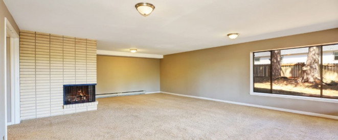 Leerstand: Darf eine Wohnung leer stehen? Ein Wohnzimmer mit einem großen Fenster, Kamin und Teppichboden, in dem keine Möbel stehen.
