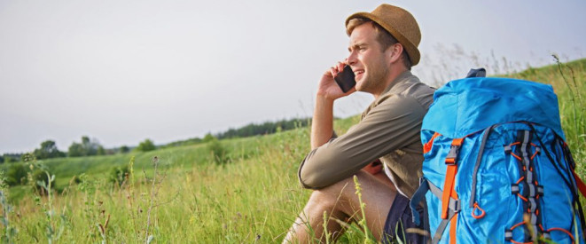 Arbeiten im Urlaub: Was ist erlaubt? Ein junger Mann mit Strohhut sitzt auf einer Wiese neben einem blauen Rucksack und telefoniert.
