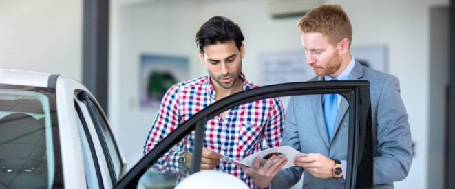 Beweislastumkehr beim Gebrauchtwagenkauf: Mehr Käuferrechte. Ein Mann im Anzug zeigt einem anderen Mann in kariertem Hemd ein Dokument. Beide stehen neben einem Auto.