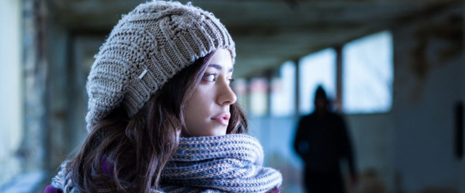 Schutz vor Stalking: Annäherungsverbot und Kontaktverbot. Eine junge Frau mit grauer Mütze und grauem Schal blickt über ihre Schulter. Im Hintergrund sieht man eine Person, die im Schatten steht.