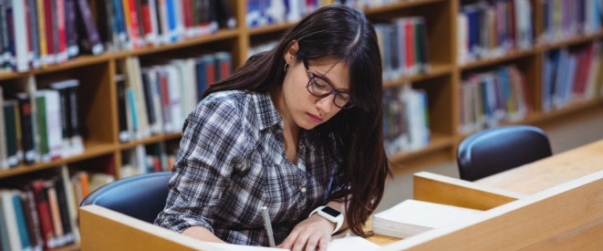 Stipendium für ein Studium: Anspruch und Voraussetzungen. Ein Mädchen sitzt in einer Bibliothek an einem Arbeitsplatz und schreibt etwas auf ein Papier.