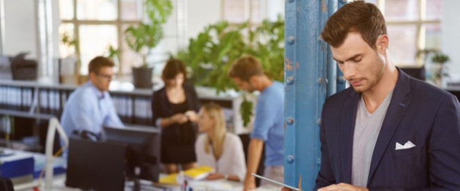 Arbeitsstättenverordnung 2016: Das sind die Neuerungen. In einem offenen Büro steht ein Mann, der auf ein Tablet schaut. Im Hintergrund stehen mehrere Personen an einem Schreibtisch.