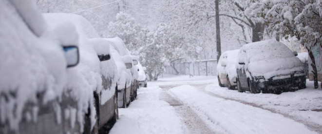 Autofahren im Winter: Gefahren und mögliche Bußgelder. Am Straßenrand einer stark verschneiten Straßen stehen mit Schnee bedeckte Autos.