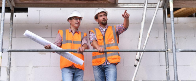 Arbeitnehmerhaftung: Was Angestellte wissen sollten. Zwei Männer in orangenen Warnwesten und Schutzhelmen stehen auf einem Gerüst vor einer unverputzten Hauswand.