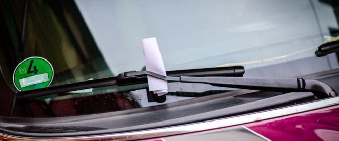 Notorisches Falschparken: Fahrerlaubnis-Entzug möglich. Hinter dem Scheibenwischer eines roten Autos klemmt ein rosa Zettel.