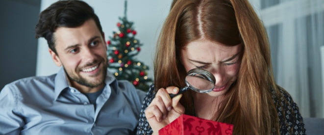 Weihnachtsgeschenke umtauschen: Das ist möglich. Ein junger Mann grinst eine junge Frau an, die mit einer Lupe ein verpacktes Geschenk betrachtet. Im Hintergrund ist ein Weihnachtsbaum zu sehen.