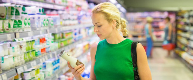 Zucker, Fett und Co.: Nährwertkennzeichnung ist Pflicht. Eine blonde Frau in grünem Kleid steht an der Kühlwarenabteilung in einem Supermarkt und hält eine kleine Milchflasche in den Händen.