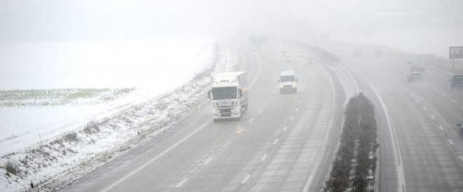 Eisplatten auf dem Lkw: Was gilt bei einem Unfall? Eine vernebelte dreispurige Autobahn, neben der Schnee liegt. Ein LKW und ein Transporter fahren auf der linken Seite.