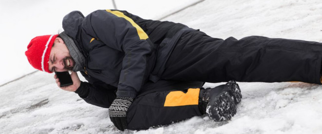 Glätteunfall auf dem Gehweg: Beweislast und Schmerzensgeld. Ein Mann liegt im Schnee, er hält sich mit deiner Hand das Knie. In der anderen Hand hält er ein Handy.