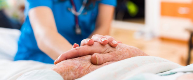 Pflegeheimkosten: Erbe ausgeschlagen – Tochter zahlt dennoch. EIne Pflegerin legt ihre Hand auf die zusammengefalteten Händer eines Seniors.