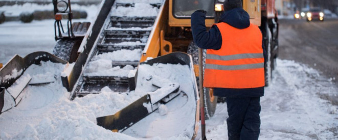 Kältefrei: Besteht ein Anspruch darauf? Ein Arbeiter mit orangener Warnweste steht, auf eine Schaufel gestützt, vor einer verschneiten Baustelle.