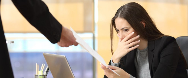 Kündigung formulieren: Darauf müssen Arbeitgeber achten. Eine Frau im Blazer sitzt am Arbeitsplatz und hält sich erschrocken die Hand vor den Mund während ihr eine andere Person ein Dokument überreicht.