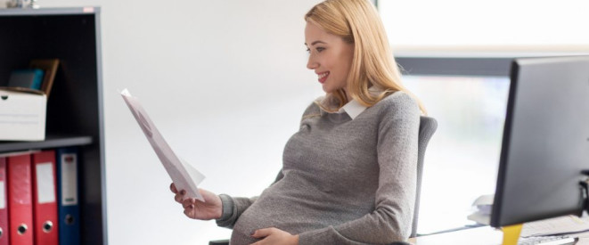 Kündigungsschutz in der Schwangerschaft: Die Rechtslage. Eine schwangere Frau sitzt an einem Arbeitsplatz und hält in der einen Hand ein Dokument und fasst sich mit der anderen Hand an den Bauch.