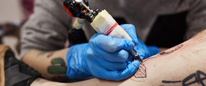 Tattoo-Fehler: Ist Schmerzensgeld möglich? Ein Tätowierer mit blauen Handschuhen tätowiert eine Männer-Wade.