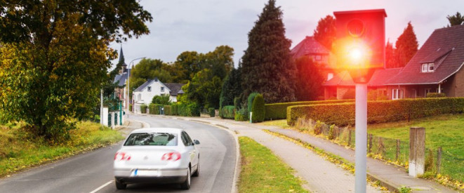 Das rote Licht einer Radarkontrolle, dahinter ein Auto auf einer Ortsstraße.