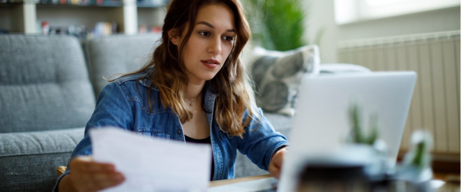 Junge Frau sitzt vor dem Sofa am Tisch und liest ein Dokument und schaut auf einen Laptop