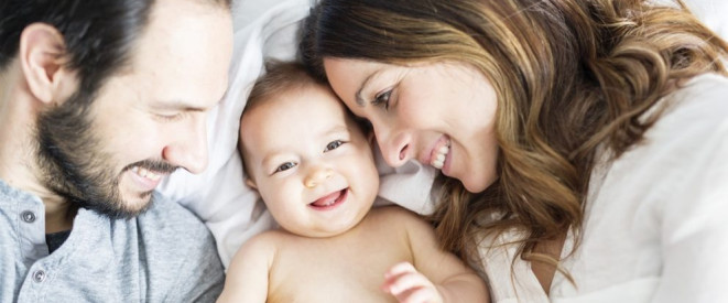 Vater, Mutter und Baby liegen lächelnd zusammen auf einem Bett.