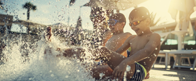 Zwei Kinder sitzen bei Sonnenschein am Beckenrand vom Pool und spritzen mit den Beinen im Wasser