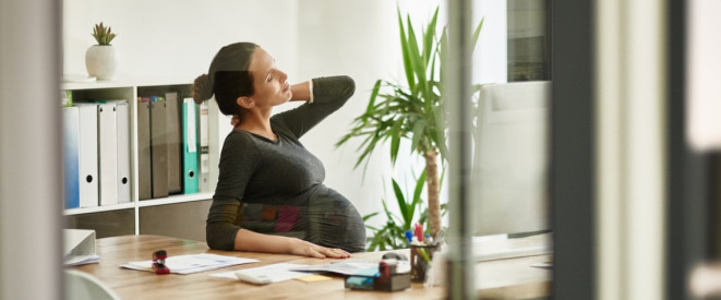 Schwangere Frau sitzt am Schreibtisch in Büro und reibt sich den Nacken