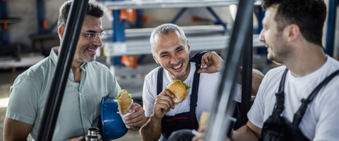 Lachende Arbeiter machen Pause in einer Fabrik und halten dabei ein Brötchen in der Hand