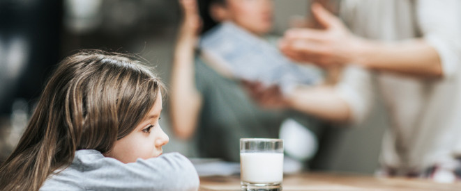 Ein Mädchen sitzt am Küchentisch, während die Eltern im Hintergrund streiten. 