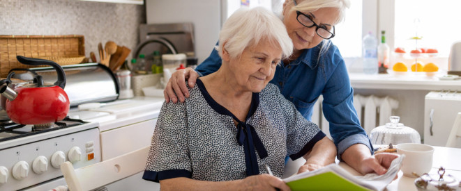 Ältere Frau unterschreibt Papiere, während jüngere Frau ihr über die Schulter schaut
