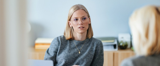 Junge blonde Frau sitzt bei einem Bewerbungsgespräch in einem Büro einer anderen Person gegenüber